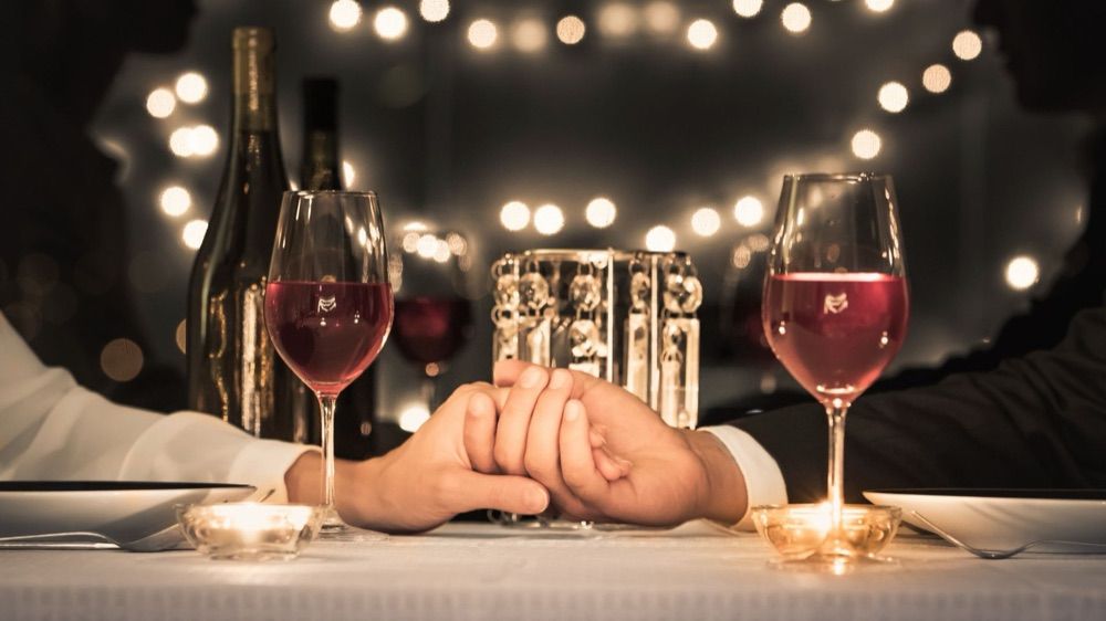 A couple's hands clasped across a candlelit table set with red wine, under warm string lights